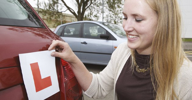 Female leaner driver handles L-plate at rear of car, day.