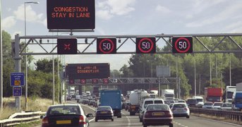 Motorway congestion speed limiter sign illuminated over heavy traffic, day.