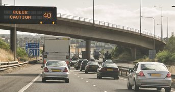 Illuminated motorway sign, queue caution, traffic passing below, day.